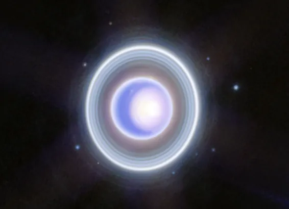 The planet Uranus on a black background. The planet appears blue with a large, white patch taking up the right half. The patch is whitest at the center, then fades into blue at it expands from right to left. A thin outline of Uranus is also white. Around the planet is a system of nested rings. The outermost ring is the brightest while the innermost ring is the faintest. Unlike Saturn’s horizontal rings, the rings of Uranus are vertical and so they appear to surround the planet in an oval shape. There are 9 blueish white dots scattered around the rings.