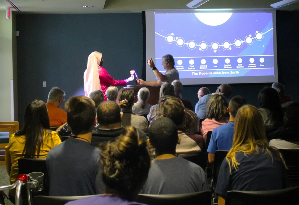 A group of people in a room with two presenters demonstrating how the Moon and Sun casts a small shadow on the Earth, resulting in an eclipse.