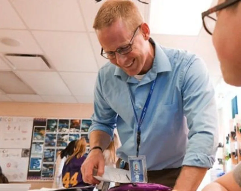 A smiling man wearing black rimmed glasses and a blue shirt with the cuffs rolled up is leaning forward, looking at something out of the frame. A young person, whose profiled face appears on the edge of the frame, is also looking at the same place. Three more young people are visible in the back of the room, standing in front of a wall of images.