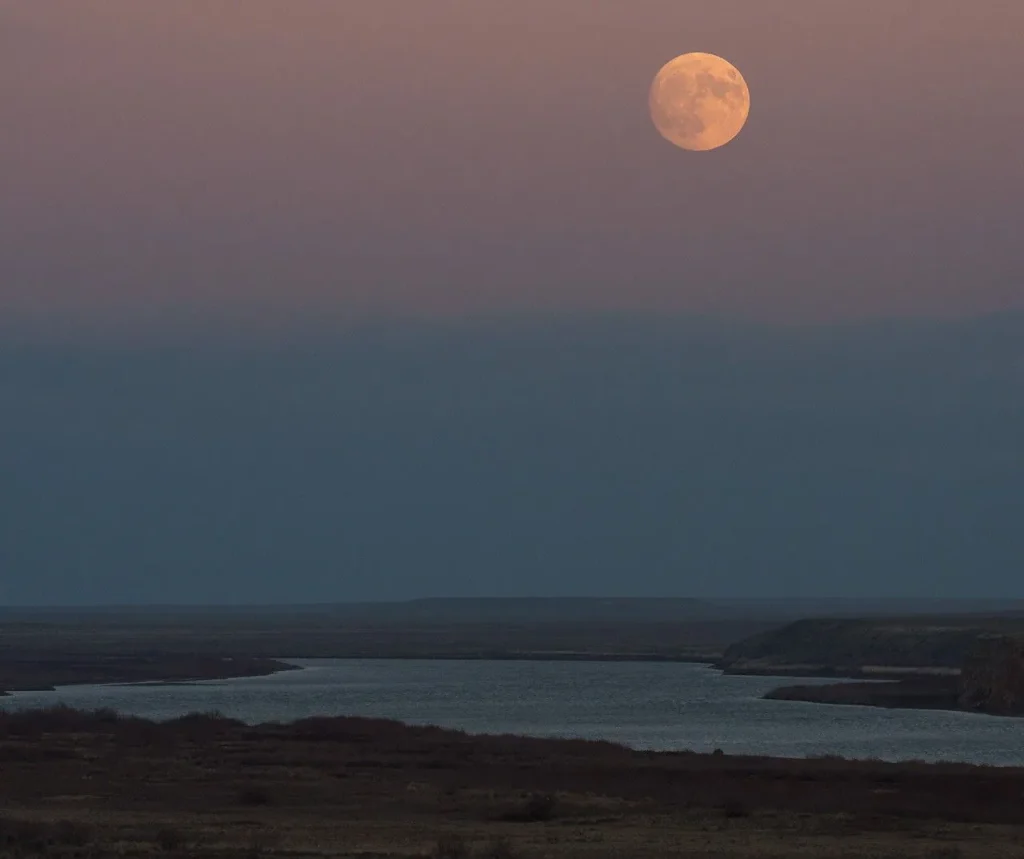 At twilight, a full moon rises over a broad river with vegetated banks. The Moon appears peach-colored in a dim pink-and-blue sky.