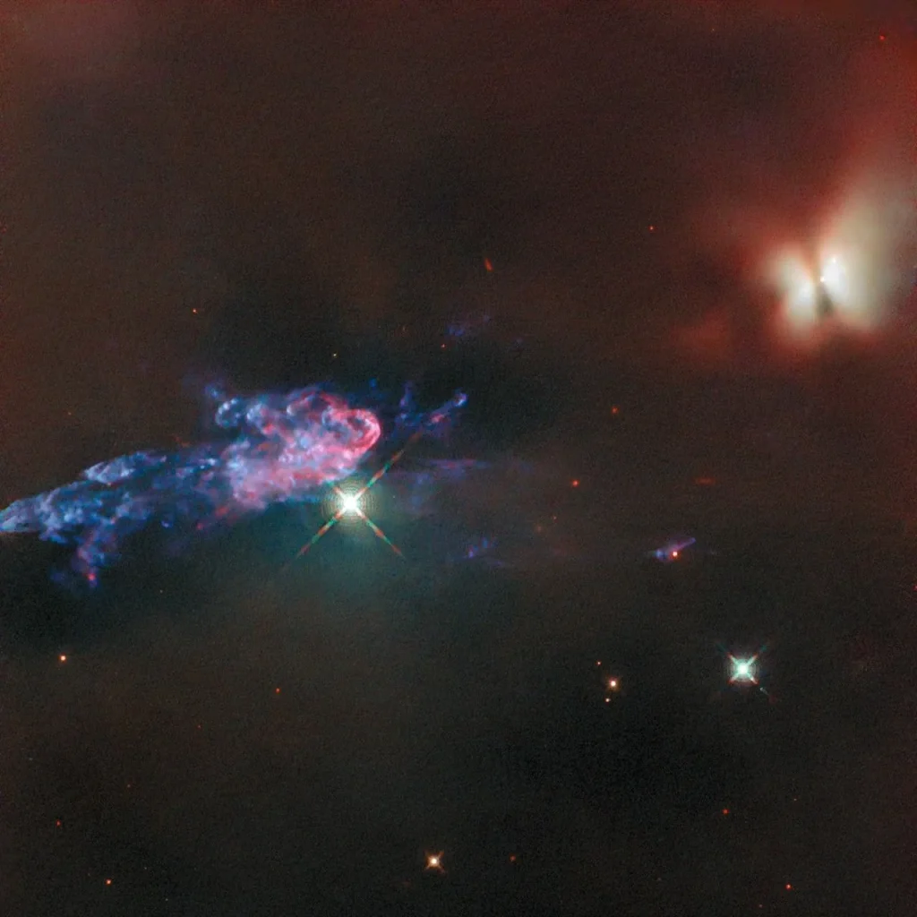 An area in the Orion nebula filled with dark, puffy clouds. On the image’s right side is a large area of clouds crossed by a dark bar. The region glows in red and whitish colors lit by a protostar within. At the other side a large jet of material ejected by the protostar appears to be made of thin, wispy, blue, and pink clouds. A couple of foreground stars shine brightly in front of the nebula.