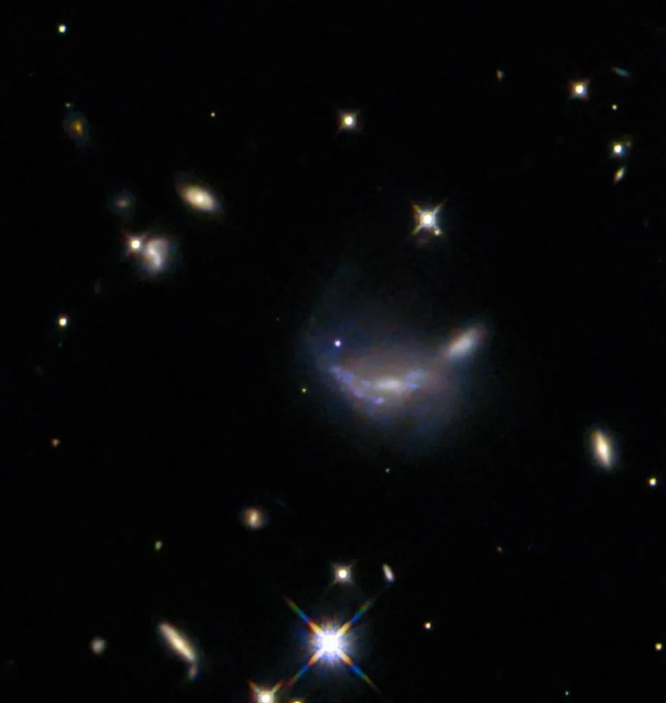In the exact center a supernova is visible as a small but bright blue dot. It lies atop the outer disk of a hazy-looking galaxy, which has a somewhat warped shape. Around this are a number of minor galaxies visible as glowing disks, and some points of light that are stars near to us, on a black background. A few bright stars hold X-shaped spikes that are optical artefacts from the telescope.