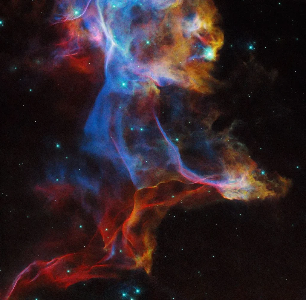 A colorful, glowing nebula that reaches beyond the top and bottom of the image. This translucent cloud of gas holds wispy and thin filaments with hard edges in some places, and puffy and opaque in others. Blue, red, and yellow colors mix together, showing light emitted by different types of atoms in the hot gas. Scattered across the colorful nebula are bright and point-like foreground and background stars. The background is black.