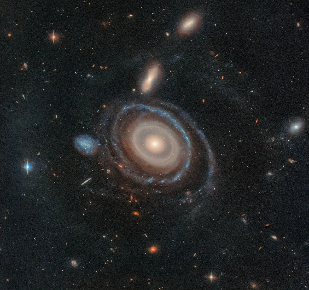 A large galaxy is at center, and a significantly smaller galaxy is to its immediate left. Both are set on the black background of space, which is dotted with a range of galaxies in different shapes and sizes, along with a few foreground stars.