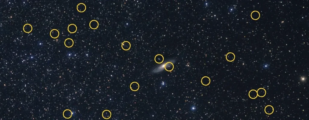 A wide-angle view of the distribution of 36 satellite galaxies orbiting the large Andromeda galaxy, which resembles bees swarming around a hive. Each dwarf is a fuzzy pinpoint identified by a yellow circle. Most of the photo is dominated by myriad pinpoint-white foreground stars inside our Milky Way galaxy.