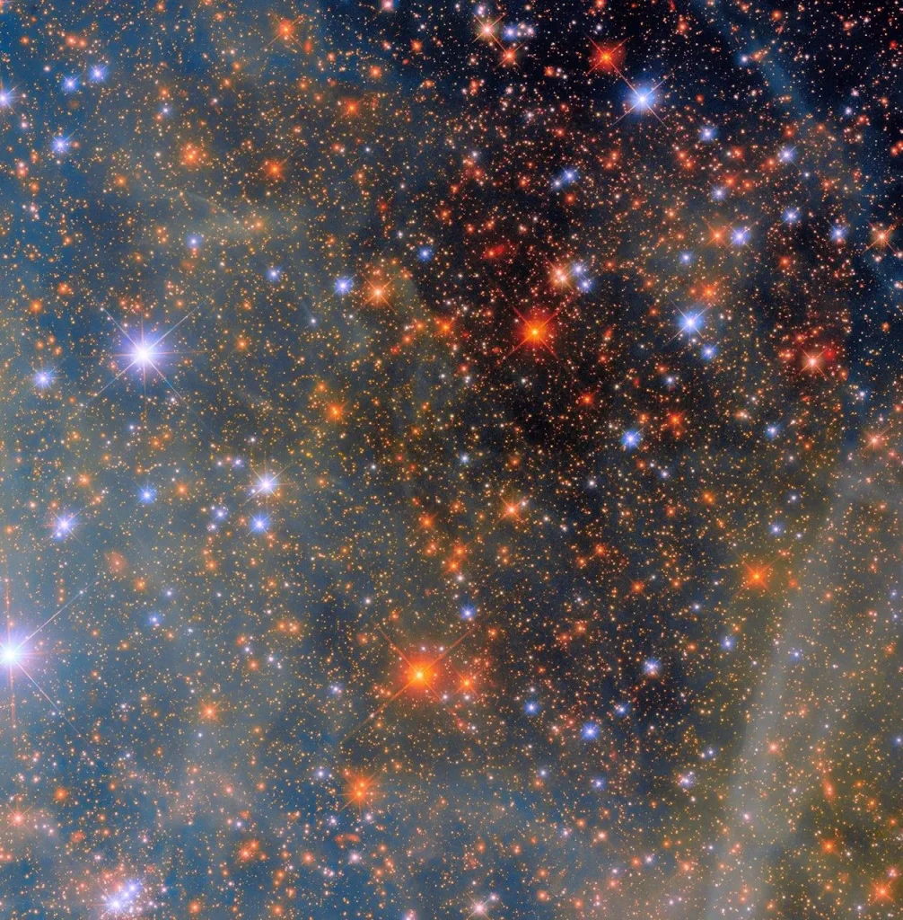 An area of space filled with stars. Most of the stars are small, distant dots in a range of orange colors; closer stars shine with a bright glow and hold four thin diffraction spikes around them. These closer stars appear both bluish and reddish. Clouds from a nebula cover the left half of the scene, giving it a blue-greenish cast. More pieces of cloud also drift over the black background of space on the right side of the image.