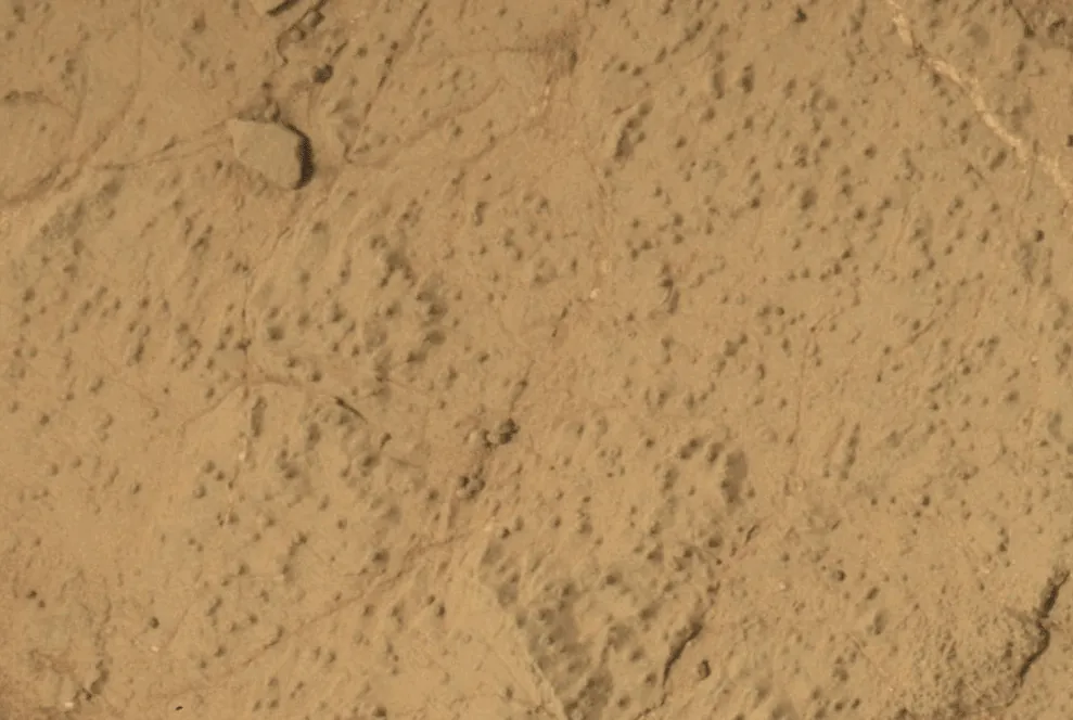 This animated image shows a close-up view of a flat, dusty, tan-colored rock surface with fine cracks and small, dark specks scattered across it. In the animation, a hole appears in the center of the frame, with loose powdered material surrounding the hole. The surrounding rock surface remains unchanged, highlighting the freshly drilled hole and the fine texture of the rock.