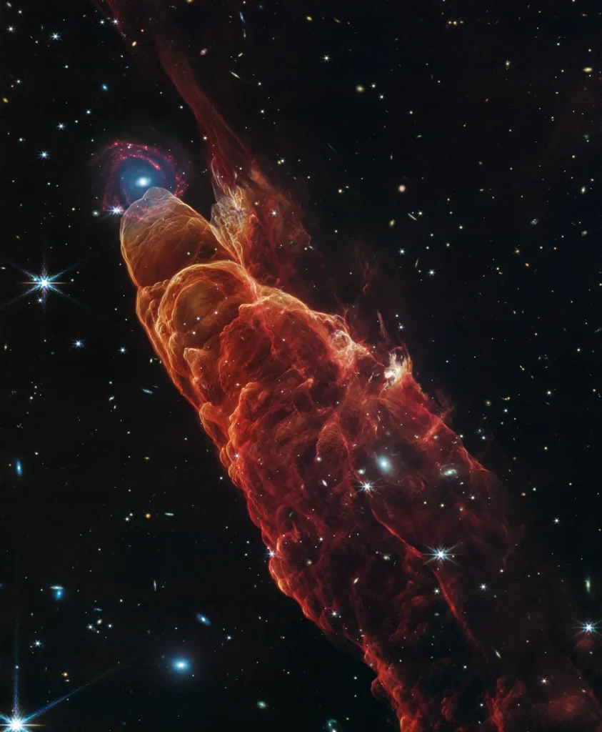 Angled from the upper left corner to the lower right corner is a cone-shaped orange-red cloud known as Herbig-Haro 49/50. This feature takes up about three-fourths of the length of this angle. The upper left end of this feature has a translucent, rounded end. The conical feature widens slightly from the rounded end at the upper right down to the lower right. Along the cone there are additional rounded edges, like edges of a wave, and intricate foamy-like details, as well as a clearer view of the black background of space. In the upper left, overlapping with the rounded end of Herbig-Haro 49/50, is a background spiral galaxy with a concentrated blue center that fades outward to blend with red spiral arms. The background of space is speckled with some white stars and smaller, more numerous, fainter white galaxies throughout.