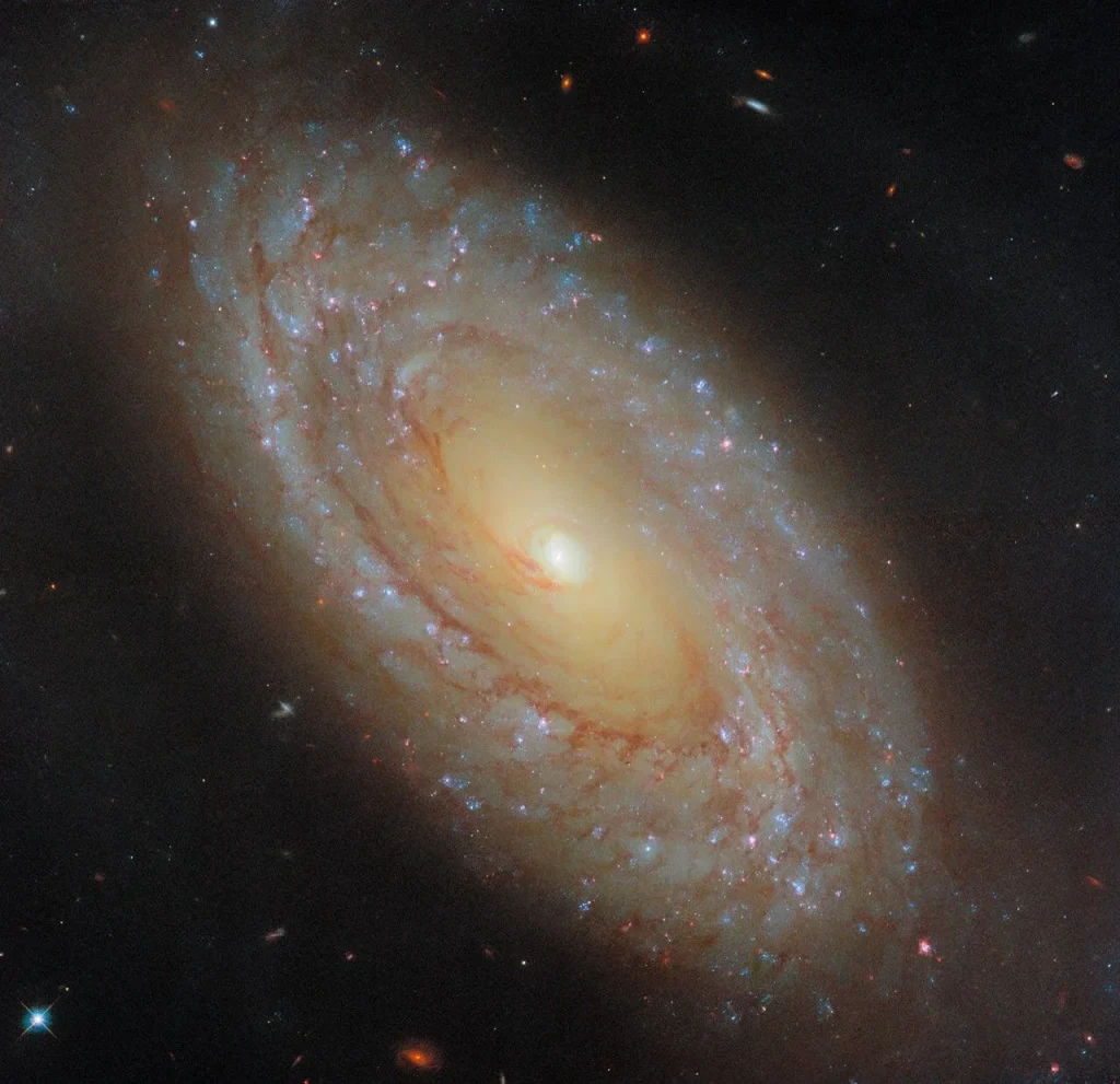 A spiral galaxy stretches diagonally across the image, from upper-left to lower-right. At its center is a bright-white, glowing orb surrounded by an inner disk of golden light that is wrapped by a broad outer disk that glows more dimly. The disk holds patchy, broken spiral arms swirling around the galaxy’s core and filled with small blue and pink star clusters. Dark, reddish threads of dust also spiral through the disk, with some strands reaching into the core.