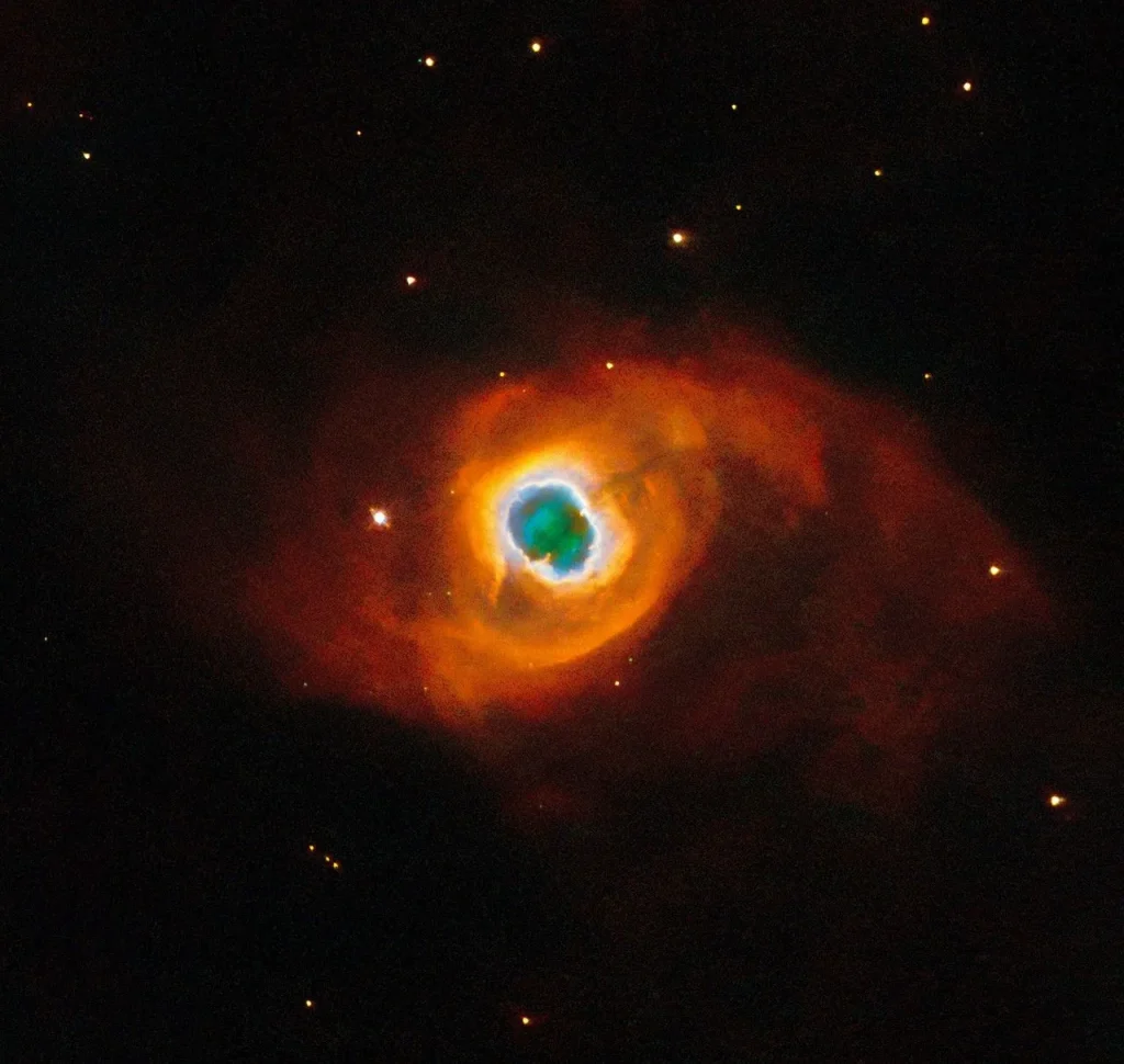 A glowing, jagged ring encircles a small central region of greenish clouds, making it appear like a hole torn in fabric. This “hole” is itself encircled by a band of silvery-blue clouds and a larger, fainter, yellow ring of gas. Puffy, smoky clouds of orange and red gas billow out from there into a large oval nebula, fading into the dark background of space dotted by stars.