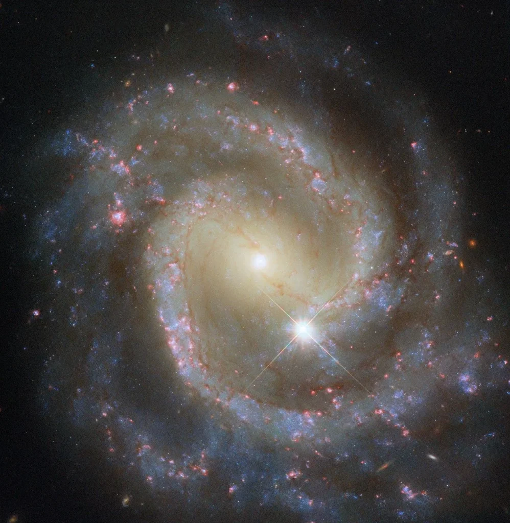 A spiral galaxy seen face-on. Across its center, a broad bar of blazing stars shines. A glowing spiral arm extends from each end of this bar, both making almost a full turn through the galaxy’s disk before fading out. The arms contain sparkling blue stars, pink spots of star formation, and dark threads of dust that follow both spiral arms into and across the central bar. A foreground star sits atop the galaxy.