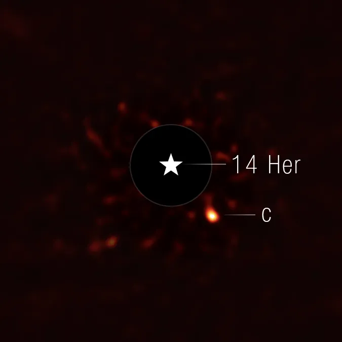 This image shows the exoplanet 14 Herculis c. The view is mostly black, with very faint red splotches in the central region of the image. At the center of the image, there is a black circle, and in the center of that, there is a star symbol representing a real star. This black circle blocks the light from the host star. To the lower right of the circle is a fuzzy bright orange circle, which is the exoplanet.