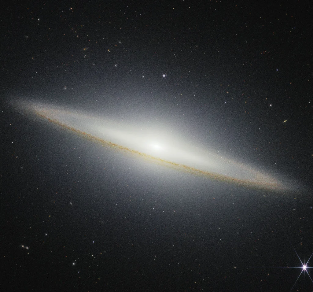 Image of a galaxy on the black background of space. The galaxy is a very oblong, brownish yellowish disk that extends from left to right at an angle (from about 10 o’clock to 5 o’clock). Mottled dark brown patches rim the edge of the disk and are particularly prominent where they cross directly in front of the galaxy. The galaxy’s center glows white and extends above and below the disk. There are different colored dots, distant galaxies, speckled among the black background of space surrounding the galaxy. At the bottom right, there is a particularly bright foreground star with Webb’s signature diffraction spikes.