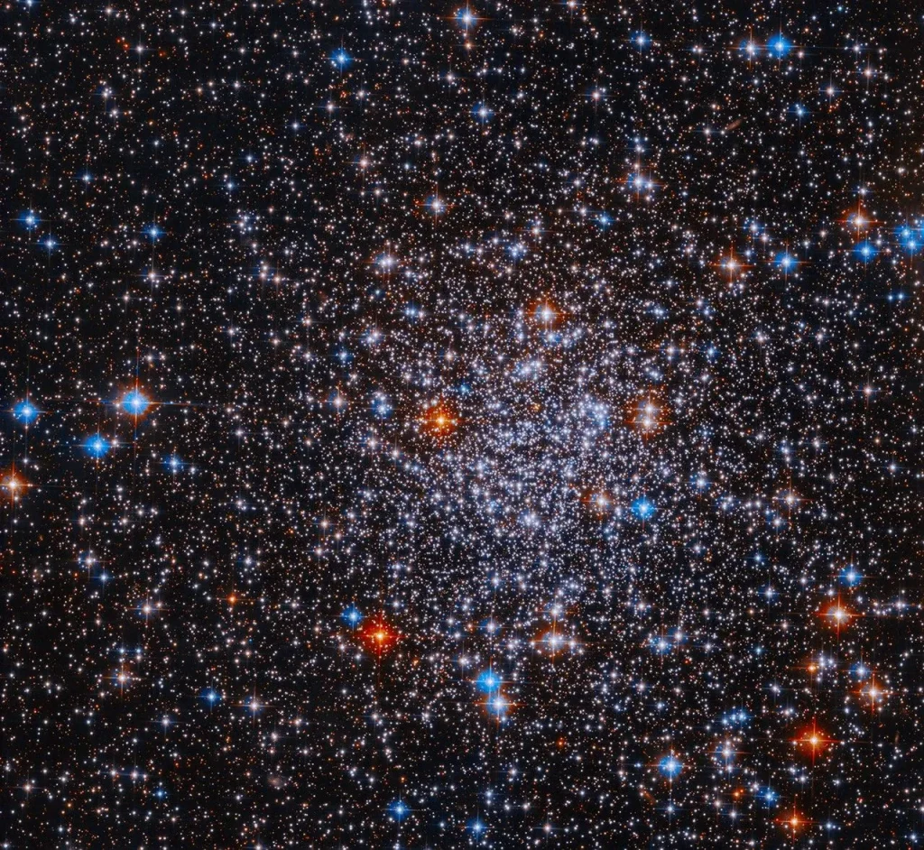 Bright stars cluster against a black background. The stars are more densely concentrated in the center of the image. The stars appear mostly white, but bright red and blue stars are also visible sprinkled throughout the image.