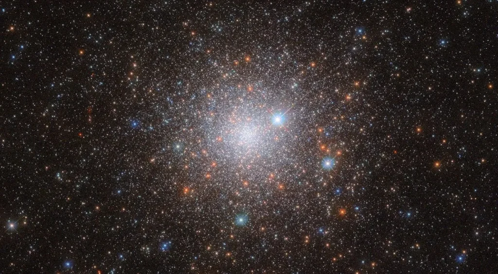 A cluster of stars in space. It’s bright in the center, where the stars are densely packed together in the cluster’s core and grows dimmer and more diffuse out to the edges, as the stars give way to the dark background of space. A few orange stars are spread across the cluster, but most are pale, bluish-white points of light. Three large stars with cross-shaped diffraction spikes around them lie between us and the cluster.