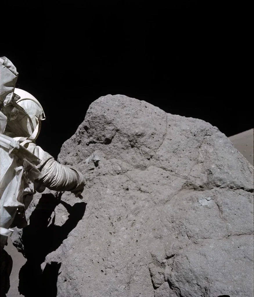 A person wearing a bulky, white spacesuit is partially visible on the left side of the image, extending a gloved hand toward a large, rough-textured, gray boulder. The boulder has a dusty, uneven surface with cracks and crevices. The person and boulder are lit up, while the background is mostly black. The person’s shadow provides a black shape cast on the boulder, highlighting the contours of both the boulder and the person.