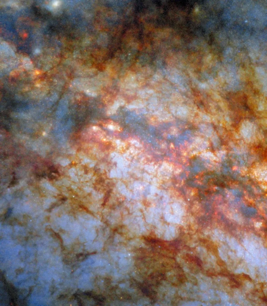 A close-in view of the center of galaxy M82. Bright, bluish light radiating from the center is due to stars actively forming there. A thick lane of gas, black in the center and red around the edges, crosses the center and blocks much of the light. Thinner strands and clumps of reddish dust cover much of the rest of the view.