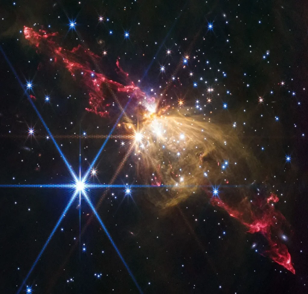 Gaseous yellow-orange filaments look like a rose seen from the side and tilted slightly from upper left to lower right, slightly higher than the center of the frame. Extending from the rose to upper left and lower right are gaseous outflows that appear as red lobes that have an overall shape of tall, narrow triangles with rounded tips. Each red triangle is made up of wavy, irregular lines. Dozens of stars are scattered across the field. One particularly bright white star with eight diffraction spikes is located at the top of the yellow rose. Another bright blue star with even more prominent diffraction spikes is to its lower left. The background of space is black.