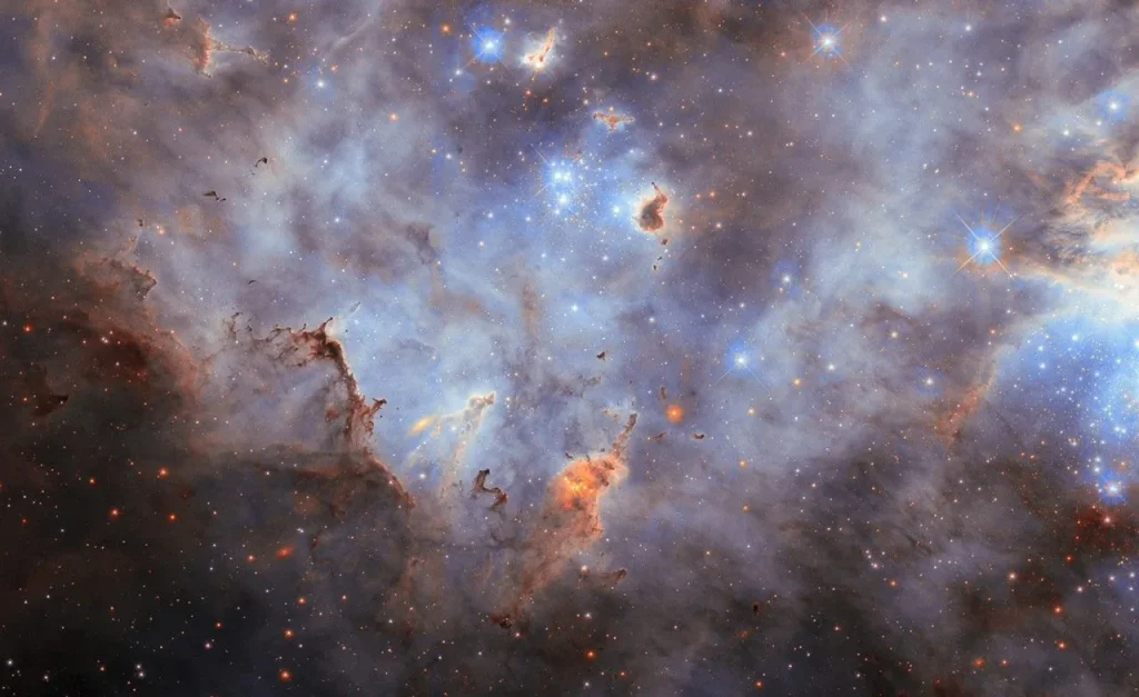 Stars in a star cluster shine brightly blue, with four-pointed diffraction spikes radiating from them. The center shows a small, crowded group of stars while a larger group is partially visible on the right side of the image. The nebula is mostly thick, smoky clouds of gas, lit up in blue tones by the stars. Clumps of dust hover before and around the stars; they are mostly dark but lit around their edges where the starlight erodes them.