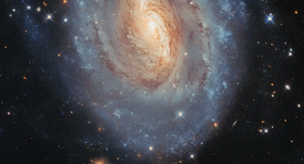 An oval-shaped spiral galaxy. Only the center and lower half of the galaxy is in frame. Its center is primarily golden in color with a white glowing core, while its thick spiral arms are mostly blue, particularly at the outskirts; these colors merge in between. Dark lanes of dust swirl through the center, blocking some of the galaxy’s light. Stars and distant galaxies are visible around the edges on a black background.