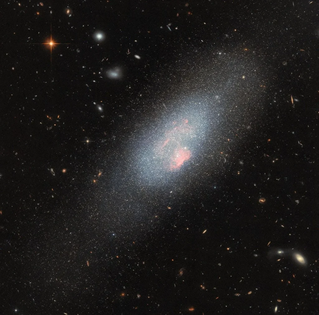 A pale blue dwarf galaxy on the black backdrop of space with some faraway galaxies. The galaxy itself resembles a fuzzy cloud of tightly packed stars, with a broad halo of stars dispersed around it. Spread across the galaxy’s core are several small, glowing patches of gas where there is a concentration of very hot stars.