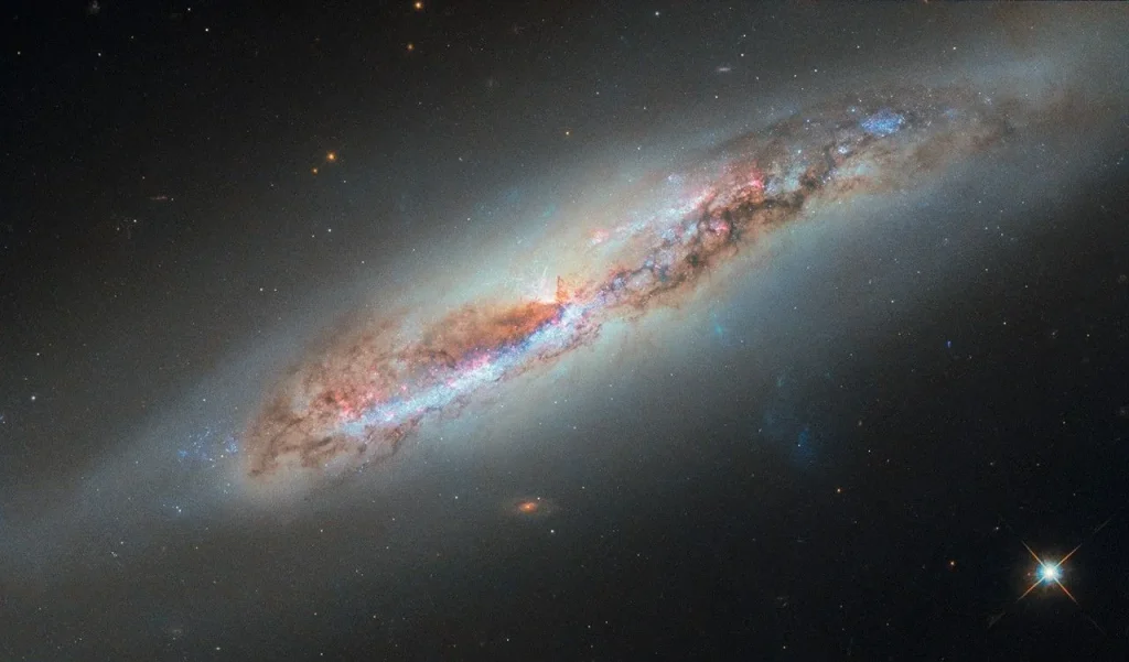 A nearly edge-on spiral galaxy. Its disk holds pink light from star-forming nebulae and blue light from clusters of hot stars. Thick dark clouds of dust block the strong white light from galaxy’s center. A faint, glowing halo of gas surrounds the disk, fading into the black background of space. A bluish plume of gas also extends from the galaxy’s core extending toward the lower-right corner of the image.