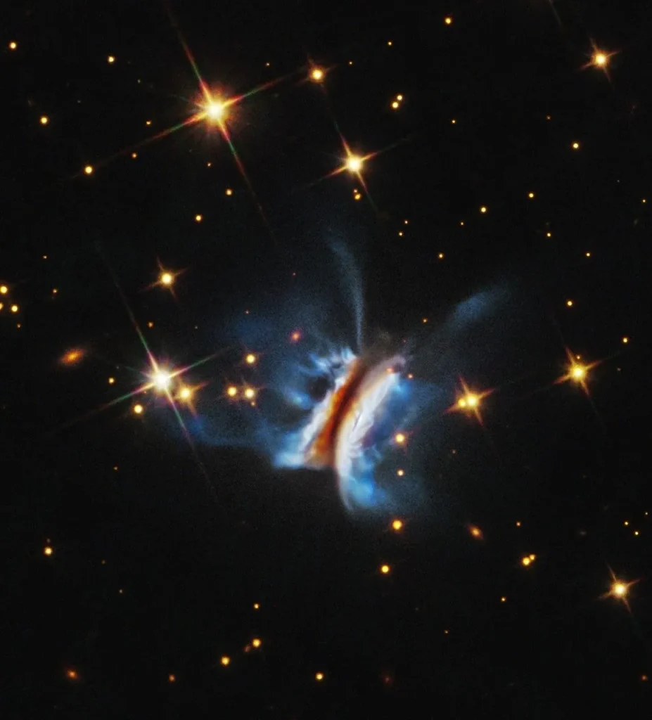 Near the center is an object that resembles an edge-on view of a hamburger. There is a diagonal dark strip (the meat patty) of dust, running from 1 o'clock to 7 o'clock, that obscures a central star. Curving away from either side of the dark strip are glowing white clouds (the buns) where dust is reflecting starlight. Bright blue finger-like wisps of material extend far above and below the dark center plane. A few dozen stars, some with four diffraction spikes, are scattered on the black background of space.