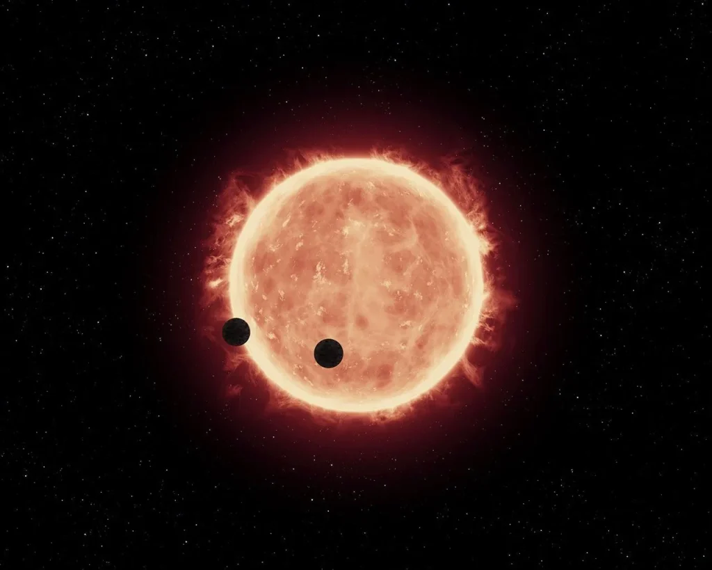A bright red-orange star burns in the center of the image, with red flames burning off it in all directions. Two dark gray spheres orbit in front of the star, one on the far left (partially in front of the star, partially off the side), and the other at the bottom center.