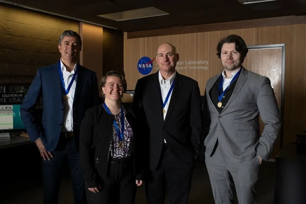 Four people standing together in a room at the Jet Propulsion Laboratory. They are all wearing suits or professional attire and have blue ribbons with gold medals around their necks. The NASA logo and the words "Jet Propulsion Laboratory" are visible on the wall behind them. A sign that says "COLD ATOM LAB" is also visible in the background.