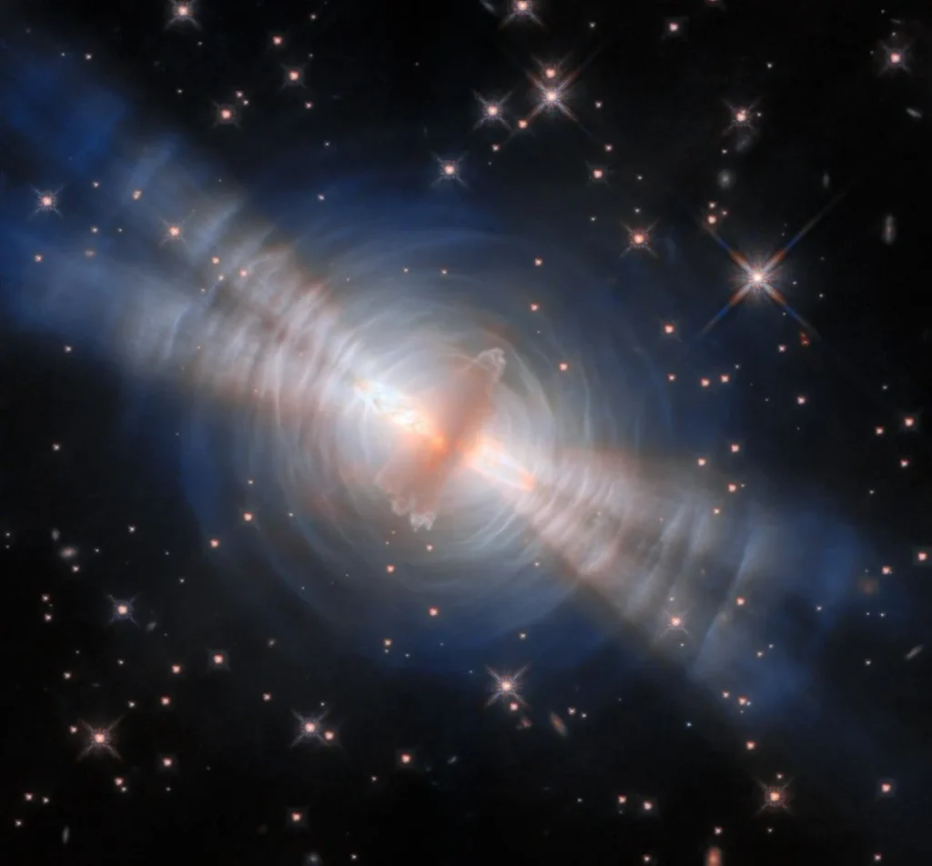 In the image center, an opaque oval cloud of gray gas aligned from 1 o’clock to 7 o’clock hides a star. Two strong beams of light from the star emerge from large holes in both sides of the cloud, forming narrow cones extending toward 10 o’clock and 4 o’clock. The central cloud is surrounded by concentric, wispy shells of gas illuminated by the star’s light. The shells reflect extra light where they’re hit by the twin beams. A crowd of smaller stars with cross-shaped spikes over them surrounds the nebula on a black background.