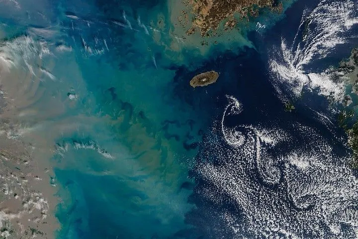 A series of spiraling clouds extends southeast from an oval-shaped island in the Korea Strait. To the west, a large sediment plume fans out from the coast of China and forms tan, teal, and blue swirls in the water.