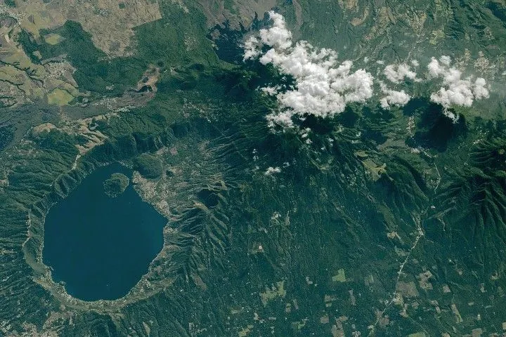 A blue lake rests within a caldera with steep walls. Several volcanoes near the caldera are capped by clouds. The terrain is mostly lush and green, with patches of gray urban areas.