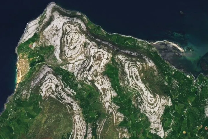 A satellite view shows green land with scattered light-brown formations containing concentric, uneven oval patterns. Blue water is visible at the top and top-left.