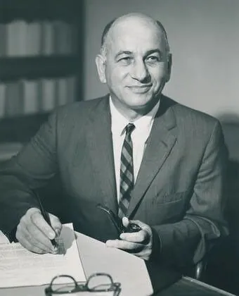 Posed portrait of William T. Pecora writing at a desk while smiling