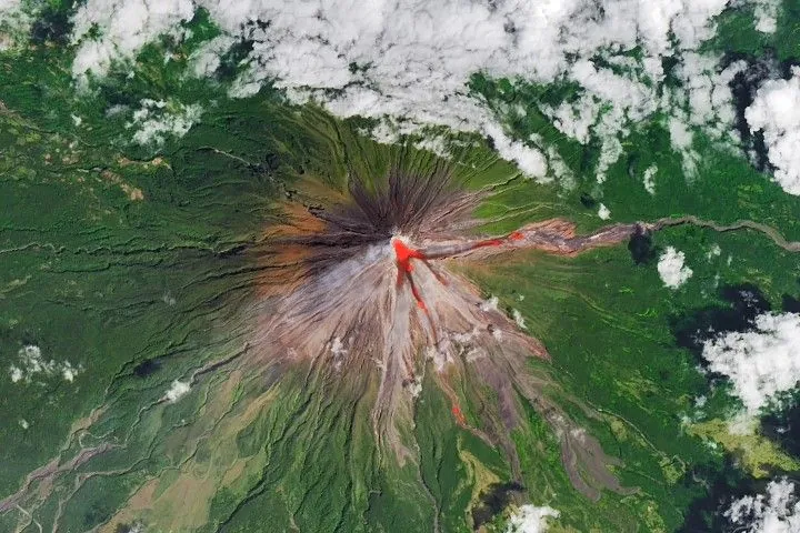 The upper slopes of Mayon volcano appear brown, with several narrow channels radiating from the crater. A red infrared heat signature appears near the summit, with red streaks extending east and southeast. The lower slopes are green and forested. Farmland and towns are visible in the lower part of the image.