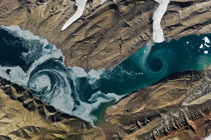 A V-shaped fjord cuts through barren brown land, with one patch of swirling water marked by white sea ice and another one colored turquoise by suspended sediment. Glacial ice flows into the fjord in several places.