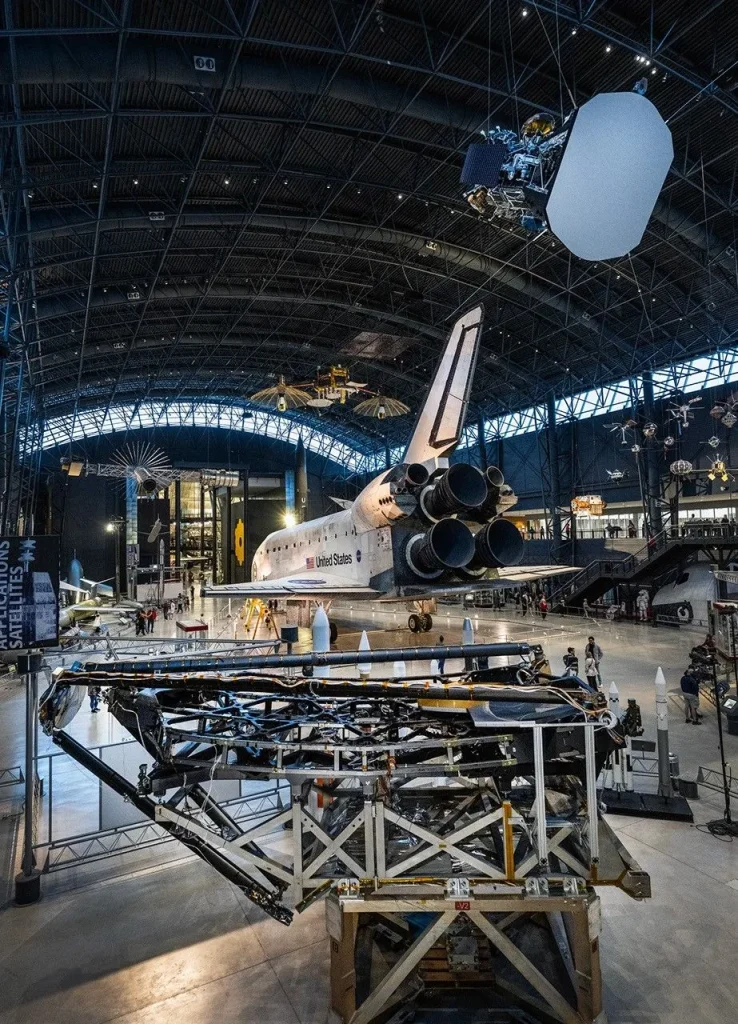 Located in a dark grey, high ceiling airplane-like hangar. A line of bright windows lines the upper part of the hangar, just below the ceiling. In the center of the image a large white and black Space Shuttle takes up most of the image, with three large conical engines in a triangular pattern on its back, with “United States” and a large American flag on its side. Hanging from the ceiling in front of the Space Shuttle is a model of the spacecraft Parker Solar Probe which has a large grey shield at its front that is curved at two ends and flat on the other two, with antenna pointing out from each of the four ends of the shield. Behind the shield a mix of shining metallic materials, and a blue square solar array with silver lines running through it. On the floor in the bottom left corner, a large metallic framework nearly 30 feet wide holds a black carbon fiber structure above it with a series of hexagonal frames on the structure. Above two of the hexagonal frames, two large hexagonal mirrors face upward, one shining in gold, and the other in silver. Above the hexagonal frame and mirrors, three large arms made of the same black carbon material rest over the top, holding a different silver circular mirror that is designed to fold out in front of the hexagonal frame, but is currently stowed. The large open room hangar has many additional smaller bright silver and gold satellites of varying size also hanging from the ceiling in the background.