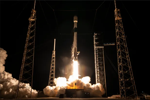Centered in the image is a rocket in the process of taking off, with bright plumes of smoke and fire emitting from the bottom of the rocket as it launches upwards. The smoke also appears in plumes on the left side of the image. Four stabilizing structures are in the foreground and background of the image, two on either side of the launching rocket. The background of the image is all black, indicating the launch took place at night.