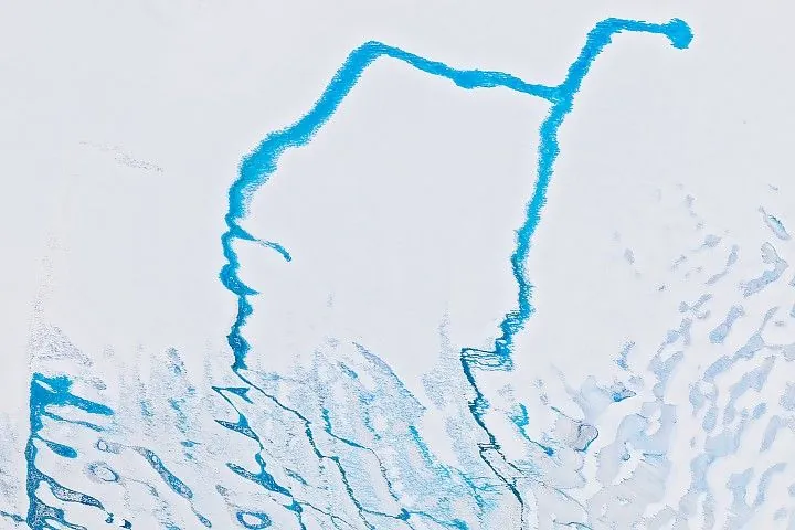 A network of cerulean blue meltwater drainage channels flowing across white and blue ice surfaces. An "oasis" of land appears as a brown rocky area in the lower part of the image.