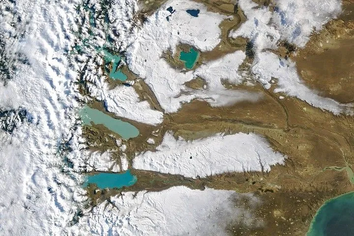 Large areas of snow cover the arid plains of southern Argentina. Several large lakes containing water in shades of milky blue are visible, while the Andes are veiled by clouds along the left side.