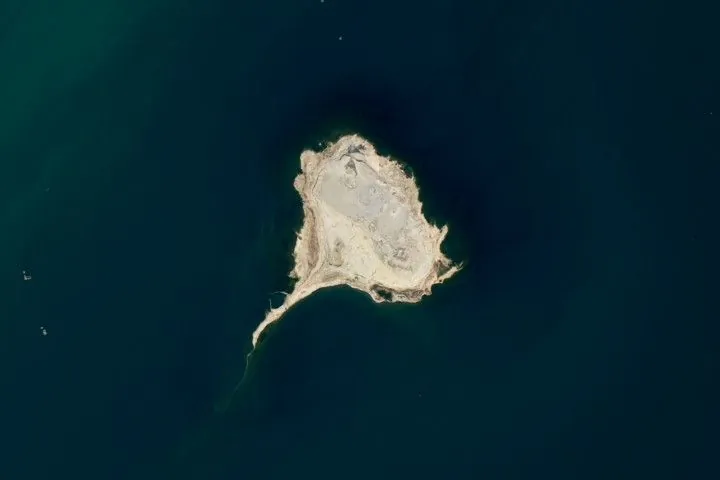 Satellite view of a tadpole-shaped brown land area encircled by blue-green water.