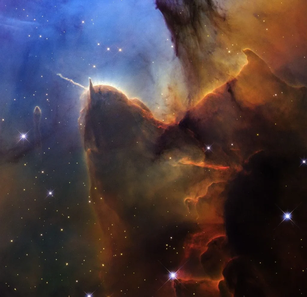 A tightly cropped Hubble view of a vast star-forming region known as the Trifid Nebula. The top left is bright blue. Brown and amber colors run from top right through the center in irregular, overlapping lines to the bottom-center. At bottom right, the view is almost black. Tiny, amber-colored stars appear throughout the scene. Toward the left there is a prominent brown shape that looks like a head with two horns. The left horn points left and is wavy. The right horn is triangular and points up. The brown dust continues, flowing down, as if along a back, and up toward the top right. A prominent line, about the same length as the left horn, appears below the middle of the body, and changes from orange to red. A small, separate semi-transparent pillar is left of the head. A few slightly larger, blue foreground stars with four diffraction spikes appear throughout.