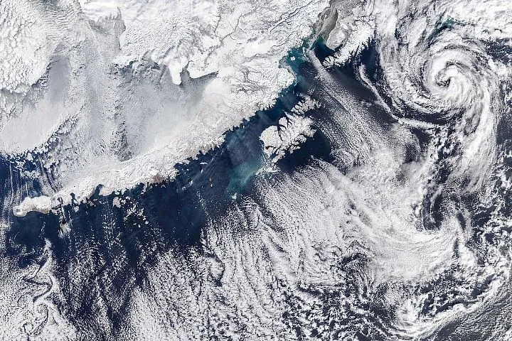 The snow-covered Alaska Peninsula is bordered by a strip of open water, which becomes obscured by varied cloud formations including vortices (bottom left), parallel cloud bands (center), and a spiral system (upper right).