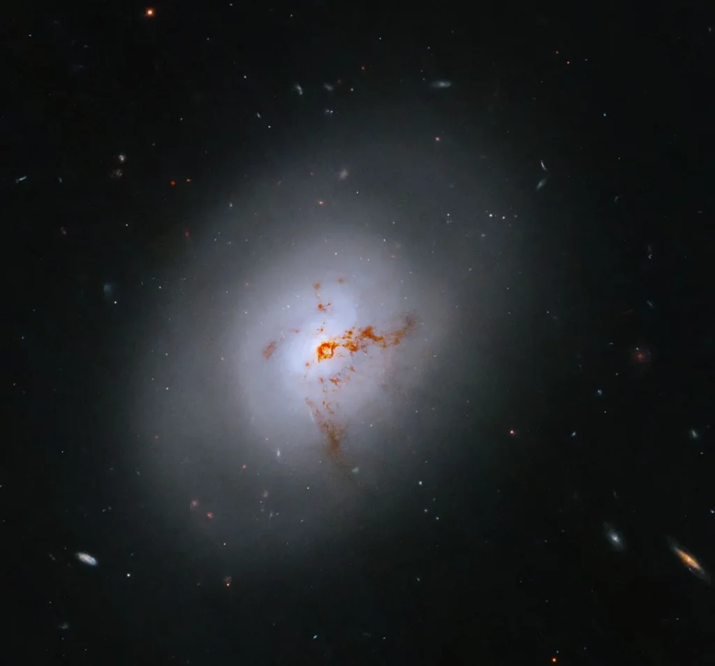 A bright lenticular galaxy seen nearly face on. Broad bright and dimmer areas of light hint at spiral structure, but there are no distinct spiral arms. Rusty-reddish-brown dust bisects the galaxy. More distant galaxies dot the black background, some even shining through the less dense regions of the bright foreground lenticular galaxy.