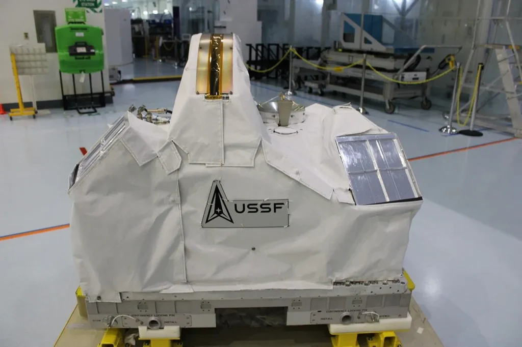 Thick, white blanketing covers irregularly shaped equipment on a pallet in the middle of a sterile-looking room with a white floor and various equipment in the background. On the blanketing is a label that says USSF. At the top of the blanketed equipment is a curved, dome-shaped section with a slit and two long gold panels running along the left and right sides of the slit. Rectangular silver panels appear on the left and right sides, and a curved, radio dish appears to the lower right of the slit.