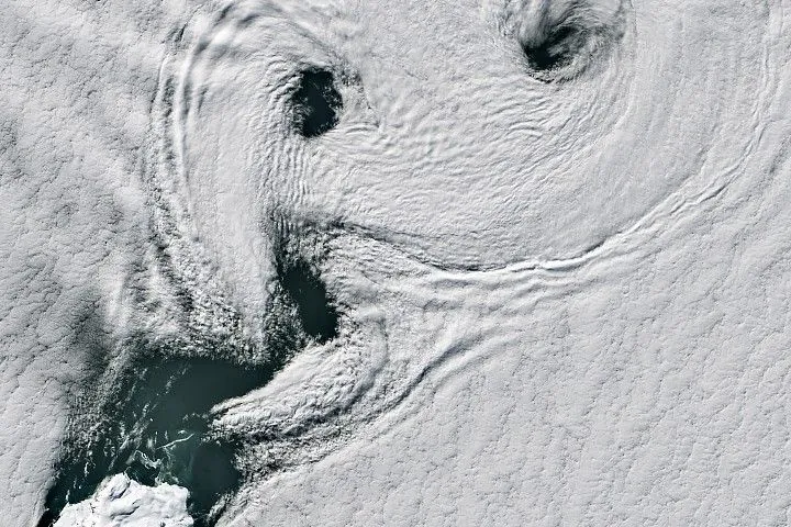 Parallel lines of cloud vortices appear downwind of a small, ice-covered island.