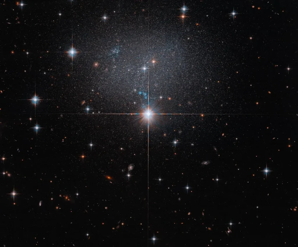 A foreground star at image center shines brightly with four diffraction spikes. Above this star is a diffuse and faint dwarf irregular galaxy. The galaxy itself has bright blue and red stars.