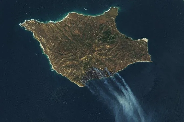 A downward-looking image of Santa Rosa Island is mostly brown, with a darker brown area on the bottom-right side. Gray-white smoke drifts toward the bottom-right over dark blue ocean water.