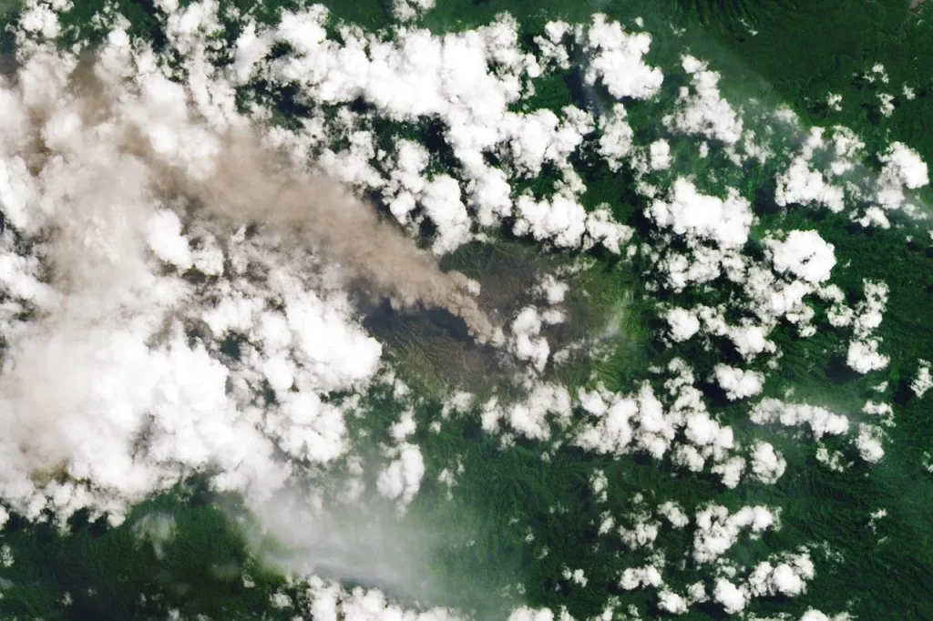 An ash-rich plume streams northwest from the volcano amidst scattered puffy, white clouds.