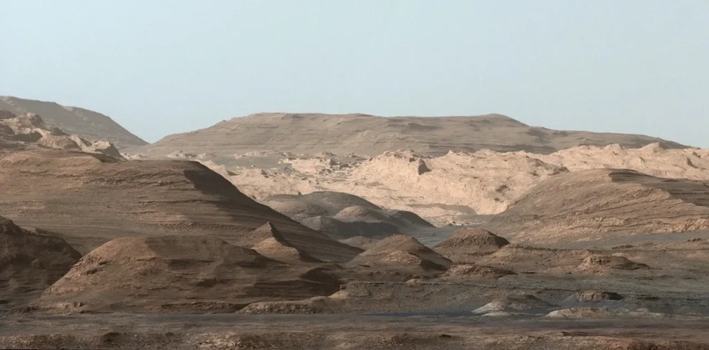 A wide-angle view of a barren, rocky Martian landscape featuring layered, wind-eroded hills and ridges in shades of tan, brown, and muted gray. In the distance, larger, more rugged formations rise toward a pale, hazy sky, showcasing the planet’s stratified geology and arid terrain.